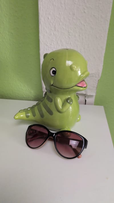 A light green, ceramic dinosaur-shaped coin bank sits on a white surface. The coin bank is approximately in the center of the frame and faces to the right, with its mouth open and tongue sticking out. It has dark green stripes on its tail and back. In front of the coin bank, a pair of black-rimmed sunglasses with rose-tinted lenses rests on the white surface. The background consists of a textured wall that is half light green and half white. The scene is indoors, with no discernible location in the city of Osnabrück, Germany, or any specific environmental features indicative of an outdoor or urban setting. There are no people, actions, or interactions visible in the image.