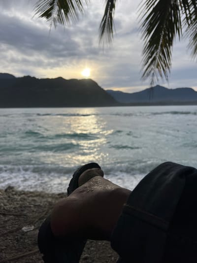 The image depicts a person's legs and feet resting on a beach, with the ocean and a mountainous landscape in the background. The sun is setting or rising, casting a warm glow on the water and clouds. Palm fronds hang from the top of the frame, framing the scene. The person is wearing dark pants and sandals, and their feet are slightly wet, suggesting they may have been in the water. The beach appears to be covered in small pebbles and debris. The location is specified as Babahie, Indonesia.