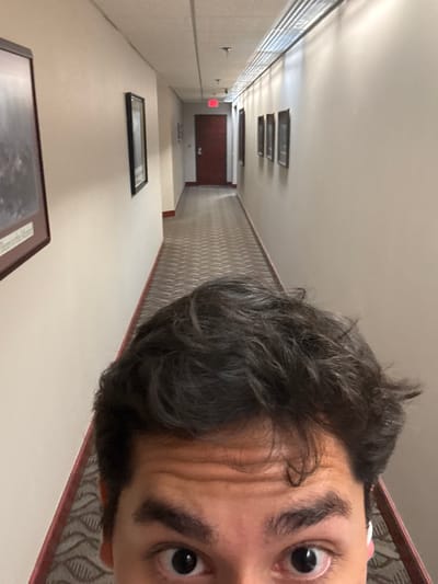 The image shows a person with dark hair and brown eyes taking a selfie in a hallway. The hallway is long and narrow with beige walls. There are framed pictures on the wall and a patterned carpet. At the end of the hallway is a closed red door with a fire exit sign above it. There are light fixtures on the ceiling. Based on the environment, the location is likely inside a building in Knoxville, Tennessee, United States of America.