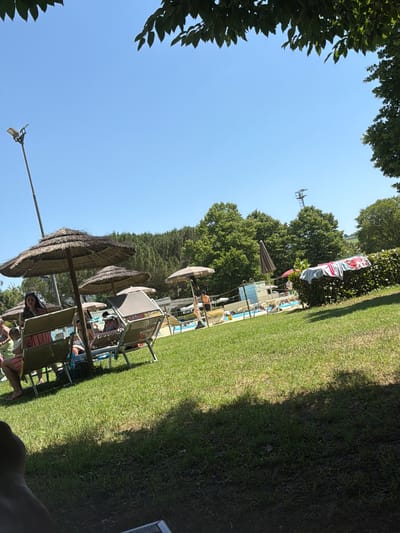 The image depicts a summer scene at a recreational area in Bertinoro, Italy. Several beach umbrellas and lounge chairs are arranged on a green lawn, where people are relaxing. In the background, there is a swimming pool with other individuals. The sky is clear and blue with trees in the upper areas, indicating a sunny day. A light post is visible among the trees.