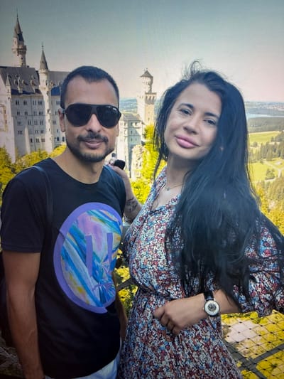 A medium shot captures a man and a woman standing outdoors with a castle and scenic landscape in the background. The man, on the left, wears sunglasses, a black t-shirt with a circular graphic, and appears to have a beard and short hair. He has a backpack strap visible on his shoulder. The woman, on the right, has long dark hair, a floral print dress, and a watch on her left wrist. Her lips are slightly parted. The background features a fairytale-like castle with towers and flags, and a hilly, green landscape that appears to be the city of Kozlovets, Bulgaria. The lighting suggests it is daytime.