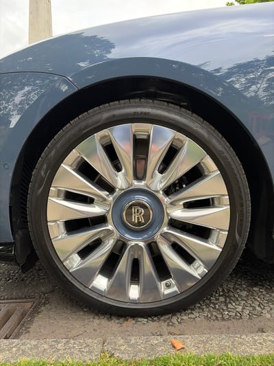 This media file presents a detailed, close-up view of the front wheel and fender of a luxury car, likely a Rolls-Royce given the distinctive "RR" logo on the hubcap. The image is taken outdoors during the daytime, under clear weather conditions, as evidenced by the bright reflections of trees and sky on the car's glossy, dark blue or grey paintwork.

The focal point is the large, multi-spoke alloy wheel, which features a highly polished or chrome finish that reflects its surroundings. The center cap is a slightly muted blue-grey, contrasting with the shiny spokes. A Pirelli P Zero tire is mounted on the wheel, displaying clear text including its size "255/40 R23 104Y", "RADIAL TUBELESS", "SEAL INSIDE", "EXTRA LOAD", and a star symbol, indicating original equipment specification. Through the wheel's spokes, parts of the brake disc and caliper are visible. A small parking sensor is also visible on the lower fender near the wheel arch.

The car is stationary, parked on a gravelly or stone-covered surface that meets a concrete curb and a strip of green grass. A metal drain grate is positioned to the lower right of the tire. A single dried leaf is visible on the grass. The setting suggests an urban or suburban environment in Harrogate, United Kingdom, emphasizing the car's presence in an everyday, yet well-maintained, outdoor space.