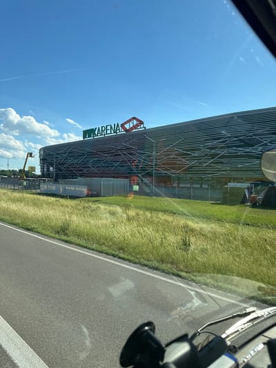 An exterior, eye-level shot captures the imposing facade of the Augsburger RFC stadium, a modern architectural marvel under a clear blue sky. The stadium's distinguishing feature is its unique, angular design, with a wave-like pattern of white metal bars crisscrossing its dark, striped exterior. Above the stadium, a large, green sign reads "ARENA" in bold letters, with a red, geometric structure framing the word.

In the foreground, a paved road with a white dividing line runs parallel to a lush, green field of tall grass that sways gently in the breeze. The field separates the road from the stadium, creating a natural buffer. Several white banners with indistinct text are erected in front of the stadium. A yellow construction lift is visible to the left of the stadium, suggesting ongoing work or maintenance. A few small figures, likely construction workers, are scattered near the stadium's entrance. The overall impression is one of a bustling construction site at a modern sports venue on a bright, sunny day.