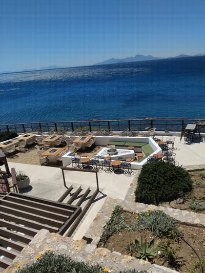 This high-angle shot captures an inviting outdoor terrace or lounge area overlooking a vast expanse of the Aegean Sea in Pyli, Greece. The scene is bathed in bright daylight, with clear skies and visible shadows indicating a sunny day, likely morning or early afternoon.

The foreground features a well-maintained garden with stone-tiered landscaping, home to various green plants, some with small yellow flowers, and a prominent dense shrub. An irrigation system is visible throughout the garden beds. Adjacent to the garden is a paved patio area.

The main subject is an outdoor dining and lounge space, meticulously arranged with various seating options. Along a protective railing that offers unobstructed sea views, multiple tables and chairs are set. These include square tables with light-toned surfaces paired with woven wicker/rattan chairs, as well as round wooden tables accompanied by dark metal chairs. A central feature is a white, built-in, L-shaped bench with light green cushions, creating a cozy lounge spot around a low table. The furniture designs suggest a modern and comfortable aesthetic.

Beyond the terrace, the deep blue sea stretches towards the horizon, where it meets a lighter blue sky. Distant, hazy mountains or landmasses are faintly visible on the horizon. The sea's surface shows subtle ripples, reflecting the sunlight.

The area appears ready for guests, but no people are present, lending a tranquil and serene atmosphere to the picturesque setting. No visible text is present in the image.