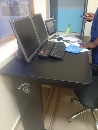 The image displays an indoor workstation located in Okengwe - Obehira, Nigeria. The central object is a dark-colored desk with a rectangular top and rounded corners. The desk's left support leg features white stenciling that reads "KSG/RHO" above a General Electric (GE) logo.
Arranged on the desk surface are three computer monitors: two identical, dark-colored flat-panel displays are positioned side-by-side on the left, with their screens off. A third, smaller, silver-framed monitor is situated to the right of the paired monitors. In front of the monitors are two black keyboards—one standard QWERTY and one specialized multi-button control keyboard—along with a black computer mouse. A green pump-dispenser bottle and a stack of papers are located behind the keyboards. A blue document or folder, featuring a white logo, rests on the desk's right side.
A partially visible individual is present on the right side of the frame. This individual is wearing a blue uniform or scrubs. Their right hand holds a dark-colored smartphone, while their left hand rests on the blue document on the desk. The individual's skin tone is dark.
The background includes a light-colored wall on the left, a window reflecting ambient light, and a blue wall section with a metallic accordion-style security gate or shutter on the right. A black office chair with a chrome base is partially visible beneath the desk. The floor directly under the desk is light-colored.
