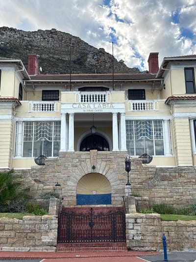 The image shows the facade of a building called "Casa Labia by Ideas Cartel" in Muizenberg, South Africa. The building has a beige exterior with white architectural details, including columns and decorative window grilles. It is made of stone and brick. A mountain rises in the background, and the sky is overcast. A portion of an ornate metal gate is visible near the entrance.