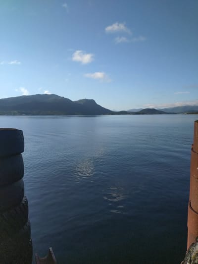 The image captures a serene natural landscape with a calm body of water stretching towards a distant mountain range. The sky is a clear blue with scattered white clouds. In the foreground, on the left side, a stack of black tires is visible, likely serving as fenders or protective barriers. On the right side, a rusty metallic structure, possibly a dock piling, is partially in view. The water's surface is slightly rippled, reflecting the soft light of the sky. The mountains in the distance are dark and silhouetted against the horizon. The scene suggests a tranquil environment, possibly a harbor or a coastal area. The location is identified as Bremsnes, Norway.