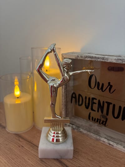 An artistic, silver-toned figurine of a woman in a yoga pose stands on a marble base. Behind the figurine, three tall, unlit pillar candles are encased in clear glass cylinders. To the right of the figurine is a rustic wooden box with a glass front. The box has the words "Our Adventure Fund" painted on it in black cursive letters. The items are arranged on a wooden surface. The image is well-lit, with a warm glow emanating from the candles. There is no indication of the location being in Osypnoi Bugor, Russia.