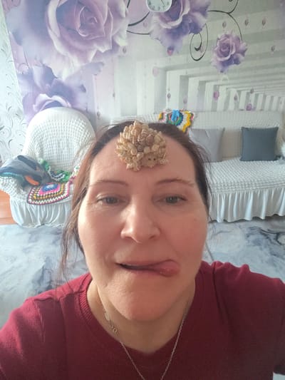 A medium-shot portrait captures a middle-aged woman indoors in Сусады-Эбалак, Russia. She looks directly at the camera with a playful expression, her tongue slightly extended to the side of her mouth. A cluster of small, light-colored, irregular objects, possibly shells or dried botanical elements, is balanced on her forehead. She has brown hair pulled back and is wearing a red long-sleeved shirt and a silver chain necklace.
The background reveals a domestic setting, likely a bedroom or living room. The wall is covered in wallpaper featuring large, stylized purple roses, green leaves, and decorative vine-like elements with small, translucent purple bead-like drops. In the upper left corner, part of a clock showing "8:" is visible, appearing reflected.
To the woman's left, a white, woven armchair is visible, adorned with a colorful crocheted blanket or throw and other fabrics. To her right, a white bed or sofa with a ruffled base is covered in a textured white bedspread. Two pillows, one light grey and one dark grey, rest on the bed/sofa. The floor is covered by a patterned rug or carpet in shades of blue, grey, and white, resembling abstract marble. The overall impression is casual and personal, with a sense of whimsy due to the woman's pose and the object on her forehead.