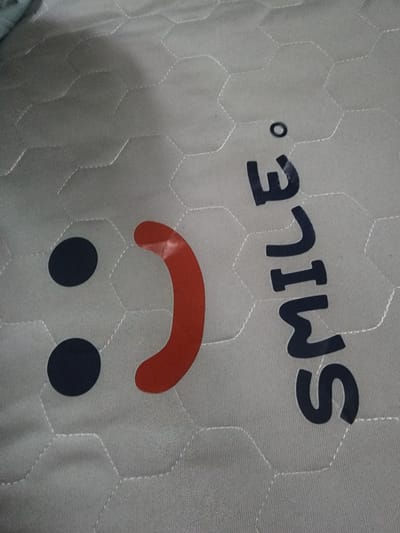 The image displays a close-up view of a light gray, hexagonal-patterned fabric, possibly a mattress cover or a blanket. A dark blue text reads "Smile." vertically in the right half of the frame, with a small black circle above it, implying a smiley face graphic. Two black circles are visible on the left, representing eyes, and a red, curved shape forms the mouth. The fabric shows stitching that follows the hexagonal pattern. There are no people or identifiable landmarks of Nonsan-si, South Korea in the image.