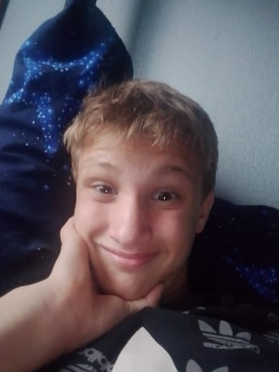 This is a close-up, selfie-style media file of a young Caucasian male. He has short, light brown, slightly messy hair and a fair complexion. He is looking directly at the camera with a wide, closed-mouth smile, his left hand cupped under his chin. His eyes are brown and wide open, conveying a cheerful and playful emotion.
He is wearing a dark t-shirt with a white Adidas logo visible on the lower right side of the frame. Behind him, to his left, is a dark blue fabric with a pattern of white specks that resemble stars or a galaxy. To his right, the background appears to be a textured, light blue wall. The lighting suggests it is daytime, possibly indoors, with a soft light illuminating his face. The overall mood is lighthearted and relaxed. No other people or objects are clearly visible. The location context provided is Kalachinsk, Russia.