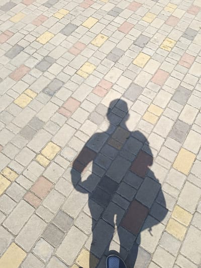 The image displays a top-down view of a paved ground surface in Maiaky, Ukraine, illuminated by direct sunlight which casts a distinct human shadow. The pavement is composed of rectangular interlocking blocks of various hues including light grey, darker grey, pale yellow, and reddish-brown, arranged in a diagonal pattern across the frame. A prominent, sharply defined shadow of a person is centered in the lower half of the image, depicting a human figure with arms bent and hands positioned on the hips. The lower-left corner of the frame shows the toe area of a blue shoe with a white sole, confirming the presence of the individual casting the shadow and taking the photograph. No other subjects or objects are discernible within the visible area.