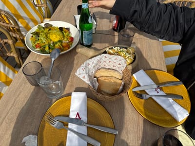 An overhead view captures a dining scene on a light-colored wooden table, likely on a restaurant terrace in Los Alcázares, Spain, illuminated by natural sunlight.
On the table, a white bowl containing a green salad with cherry tomatoes and a central yellow element, possibly a poached egg or a large dollop of dressing, is positioned centrally-left with a spoon resting inside. To its right, a green glass bottle of S.PELLEGRINO sparkling water stands, with a person's right hand, wearing a black long-sleeved jacket, reaching towards its cap from the right side of the frame. Adjacent to the bottle, a small black pan holds several potato portions topped with a white sauce and green herbs.
A woven basket containing multiple bread rolls, lined with white paper featuring a repeating red "A" pattern, is situated below the potatoes. Two distinct place settings are visible: one on the left with a round yellow plate, a white napkin embroidered with a red "A", and a silver fork and knife. Another similar place setting is partially visible on the lower right, also with a yellow plate, white napkin, and cutlery.
Further to the left, two clear glasses are present; one is ribbed and empty, while the other contains ice and water. A dark cylindrical bottle is partially visible behind the S.PELLEGRINO bottle. In the background, wicker chairs with yellow and white striped cushions are visible, indicating an outdoor or semi-outdoor dining environment. Shadows are cast across the table, consistent with bright daylight.