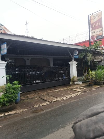 The image depicts an urban street scene. While requested to be located in Masaiti, Zambia, visual evidence within the image, including Indonesian text on signage ("DJARUM COKLAT," "TOKO TALANG," "AQUA Sumber Air Minum") and a distinct Indonesian-style vehicle license plate, indicates an Indonesian location.
The foreground reveals a partial view of a grey object, likely an interior car component. An asphalt road, exhibiting minor puddles, runs parallel to a cracked concrete sidewalk. A black metal fence forms the boundary of a property, behind which a dark-colored car with a license plate (e.g., "DK 74xx xx") is partially visible. The property features a single-story building with multiple dark, closed roller shutters and a prominent white architectural pillar supporting a blue and white "AQUA" sign. Green potted plants are arranged on the sidewalk to the left, one contained within a tire. To the right, a tall vertical billboard displays "DJARUM COKLAT," "ELITE," and "TOKO TALANG." Palm-like trees and additional potted plants are situated on the right side of the property. The sky is overcast. A communication tower is visible in the background on the left. No individuals or active interactions are discernible within the scene.