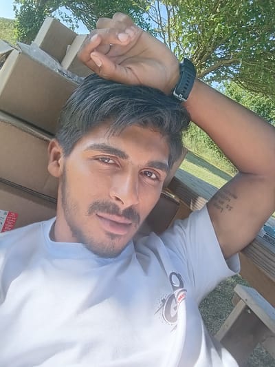 The image shows a man lying down, looking at the camera. He is wearing a white t-shirt with a graphic logo on it. His head is resting on some cardboard boxes, which are stacked behind him. The scene appears to be outdoors, with green grass and trees visible in the background. The sky is blue and appears to be a clear day. The man has a beard and mustache and his hair is dark. There is a tattoo on his left arm that reads "Love is an open not only". He is wearing a black digital watch on his left wrist. The photo seems to be taken during the daytime, likely a sunny afternoon. The man's expression is neutral. The overall setting suggests a casual outdoor environment, possibly a backyard or a park.