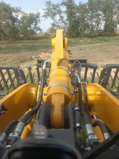 The image, taken from the perspective of a driver inside a yellow loader, shows the machinery's front hydraulic arm and attachment. The loader is situated outdoors on a dirt and grass terrain, likely in a rural area, with trees visible in the background under a clear sky. Several black tire rings and a pile of hay or straw are present on the ground. The attachment appears to be a rake or grapple, made of black metal bars. The perspective is forward-facing, with the focus on the yellow hydraulic cylinders, hoses, and metal framework of the loader. There are no people visible in the image. The location is identified as Zarinsk, Russia.