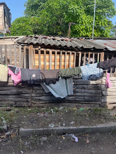The image depicts a clothesline strung across a rustic wooden fence in Moratuwa, Sri Lanka. Various items of clothing, including shirts, shorts, and what appear to be underwear, are pinned to the line with clothespins. The clothing is of different colors and patterns, and some pieces are partially obscured by others. Behind the clothesline, a wooden structure with a corrugated iron roof is visible. The roof appears weathered and rusty. Lush green trees and foliage frame the upper portion of the image, with a clear blue sky overhead. The ground in front of the fence is dirt and grass, with some scattered debris. The scene suggests a residential area, likely a home in Moratuwa, where laundry is being air-dried.