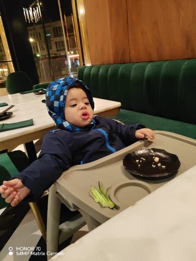 A baby in a blue hooded jacket sits in a highchair with a gray tray. A small, dark plate holds food remnants. Sliced cucumber decorates the tray. The baby has food around their mouth. The background reveals a restaurant setting with green cushioned seating and a table with dark green placemats. A window behind the baby shows building lights.