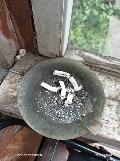An ashtray containing several cigarette butts and ash sits on a wooden windowsill. Outside the window, greenery is visible. The overall scene suggests an outdoor or semi-outdoor environment. The presence of the ashtray with used cigarettes indicates a recent or ongoing smoking activity. The wooden windowsill appears weathered and aged, suggesting it may be part of an older structure. The lighting suggests it is daytime. There are no people visible in the image. There are no indications that this scene is located in Akhalkalaki, Georgia.