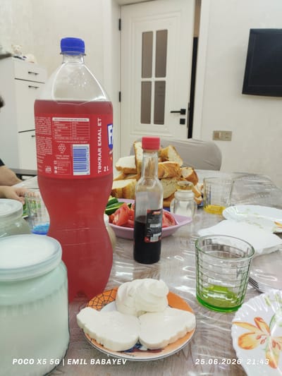 This media file captures a close-up view of a table laden with food and drinks, likely a meal setting in Azerbaijan. The focal point is a large bottle of red soda with a blue cap, bearing text in Azerbaijani, including "TƏKRAR EMAL ET" (Recycle It). Adjacent to it is a small glass bottle with a red cap, containing a dark liquid, possibly a condiment like vinegar or soy sauce, with a label indicating it's from "Secma".

The table is covered with a transparent plastic tablecloth. A variety of food items are visible: sliced bread piled high, a plate with white cheese that appears soft and possibly fresh, and a pink dish containing chopped tomatoes and cucumber slices. There are also several glasses, one of which contains a green liquid, possibly juice or a beverage, and another with a yellowish liquid. In the foreground, a large, opaque white jar with a light blue lid is present, its contents unclear but appearing to be a dairy product or liquid.

In the background, a portion of a door with glass panes and a black handle suggests an indoor setting, possibly a home or restaurant. A piece of electronic equipment, likely a TV, is mounted on a white wall. A glimpse of a person's arm and hand can be seen on the left side of the frame.

The timestamp "25.06.2026 21:43" and the text "POCO X5 5G | EMIL BABAYEV" indicate the time and the device used for capture. The presence of various food items and drinks suggests a meal is either in progress or about to begin. The setting appears casual and domestic. The lighting suggests it's evening.