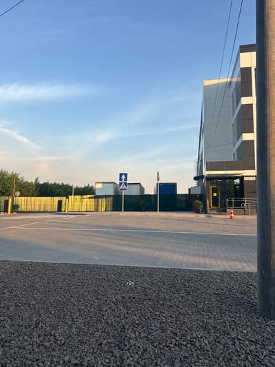 The image depicts an outdoor scene with a modern building in the background, a paved area with white markings, and a gravel-covered foreground. The building is tall and features a contemporary design with dark and white panels. Adjacent to the building, there is an entrance with a yellow door and a ramp. To the left of the building, a green fence separates the property from a road or parking area, which has a pedestrian crossing marked with white stripes. Several large shipping containers are visible behind the fence, along with trees and a red and white striped pole. A street sign showing a pedestrian and an upward arrow is posted on the right side of the paved area. The sky is blue with some wispy clouds. The overall scene appears to be an industrial or commercial area. The location is identified as Horenychi, Ukraine. There are no people visible in the image.