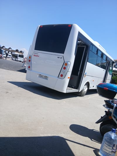 A white Isuzu HC 45 bus is parked on a paved surface under a clear blue sky. The bus is angled, with its rear facing the camera. The rear door is open, revealing a dark interior. The bus has a license plate that reads "20 691 ZAA". In the background, other vehicles, including a white minivan, are visible. Several people are standing near the vehicles. To the right of the bus, a blue and red storage box is mounted on the back of a motorcycle. A large plastic bottle of water is on the ground near the motorcycle. The scene appears to be in Gulistan, Uzbekistan, likely at a transportation hub or rest stop.