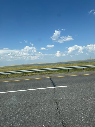This image depicts a roadside scene under a bright blue sky with scattered white clouds. The foreground is an asphalt road with a white line and a yellow line indicating lanes. A metal guardrail separates the road from a grassy embankment. Beyond the embankment is a body of water, possibly a lake or reservoir, with a treeline and a distant tower visible on the horizon. The landscape suggests a rural or semi-rural environment, consistent with the outskirts of Алтынды ауылдық округі, Kazakhstan. There are no people or animals visible in the frame, and the scene is static. The lighting suggests it is daytime, likely midday given the brightness.