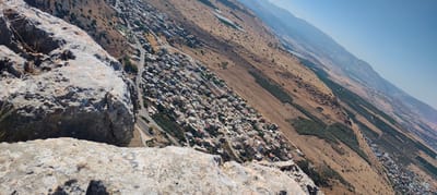 The image captures a high-angle, wide shot of a hillside town in the Galil Tachton Regional Council, Israel. In the foreground, a large, textured grey rock formation occupies the left side of the frame, obscuring the bottom left corner. Beyond the rock, the town unfolds across the slope, characterized by a dense collection of residential buildings with red-tiled roofs and white walls. A winding road cuts through the settlement, with a few vehicles visible.
The town is nestled within a landscape of arid, brown hills dotted with patches of green vegetation, likely agricultural fields or orchards. These fields are arranged in neat rows, suggesting a cultivated area. The terrain slopes downwards towards the right and recedes into the distance, where hazy blue mountains form a backdrop against a clear, bright sky. The overall scene suggests a sunny day, likely midday or early afternoon, given the strong sunlight and the lack of long shadows.
The perspective from the rocky vantage point gives a sense of scale and emphasizes the dramatic topography of the region. The environment appears dry and sun-baked, typical of a Mediterranean climate. There is no visible text in the image. The scene conveys a sense of peaceful coexistence between human settlement and the natural landscape, with the town integrated into the contours of the hill.