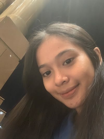 A tilted, close-up selfie features a young woman, likely taken at night or in a dimly lit indoor environment in Samarinda, Indonesia. The woman's face and upper torso dominate the frame, angled upwards and to the right. She has long, dark hair that cascades down, partially obscuring her left side and extending towards the bottom and right.

Her dark eyes are directed towards the camera, accompanied by a subtle smile. Her facial features are softly illuminated, contrasting with the predominantly dark background. One ear is visible near the top edge of the frame. In the background, indistinct structural elements can be discerned: a light-colored, cylindrical object (possibly a pillar) on the far left, and some brown, rectangular shapes (perhaps boxes or building components) in the lower left quadrant. The scene suggests a casual, personal moment captured with limited light. No text is visible.