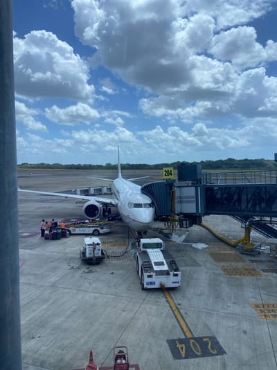 The image depicts an active airport tarmac scene at gate 204 of Tocumen International Airport, Panama. A white passenger jet, likely a Boeing 737 variant, is parked with its nose connected to a dark-colored jet bridge.

Three to four ground crew personnel are visible near the aircraft's left wing and fuselage, wearing high-visibility orange and yellow vests over light shirts and dark trousers, actively engaged in baggage handling with a white baggage loader/conveyor positioned beneath the wing. Numerous pieces of luggage are on the loader.

Various ground support equipment is present: a small white service vehicle marked "GP 27" is situated near the aircraft's front fuselage, and a large white pushback tug, marked "PB 20" and "TUG," is parked on a yellow line in the foreground, facing away from the aircraft. Yellow service hoses or cables extend from the jet bridge to the aircraft's underside.

The concrete tarmac features prominent yellow markings, including "204" in the foreground and other alphanumeric codes such as "B777" and "A3X" further back. In the background, beyond the tarmac, a dense strip of green vegetation borders what appears to be a distant runway or taxiway. The sky is bright blue with numerous scattered white cumulus clouds. The image is captured from an elevated interior perspective, likely through a terminal window, evidenced by a partial grey structural pillar on the left.