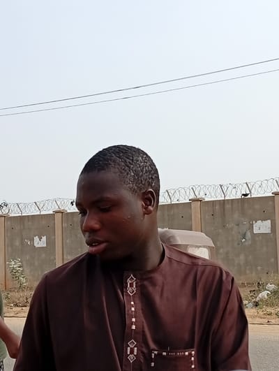 A person wearing a brown shirt with patterned embroidery is standing in front of a beige concrete wall topped with barbed wire. A vehicle is parked behind the person. The sky is overcast and a few trees are visible in the background. The person is looking slightly down and to the left. A bit of greenery is on the left side of the image.