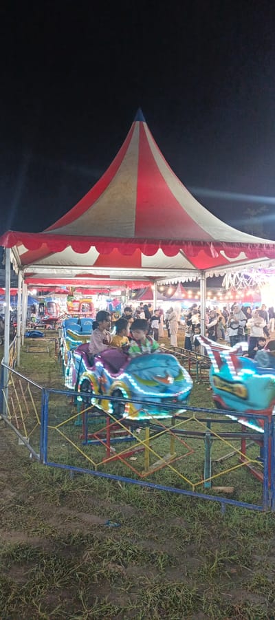 An outdoor amusement ride, likely a kiddie train or car ride, is in motion at night. The ride features several colorful, illuminated cars. Three children are seated in the foreground cars. In the background, a crowd of people are visible under a large circus-style tent with red and white stripes. The ground is covered in grass. The scene appears to be from a local fair or carnival in Sinar Semendo, Indonesia.