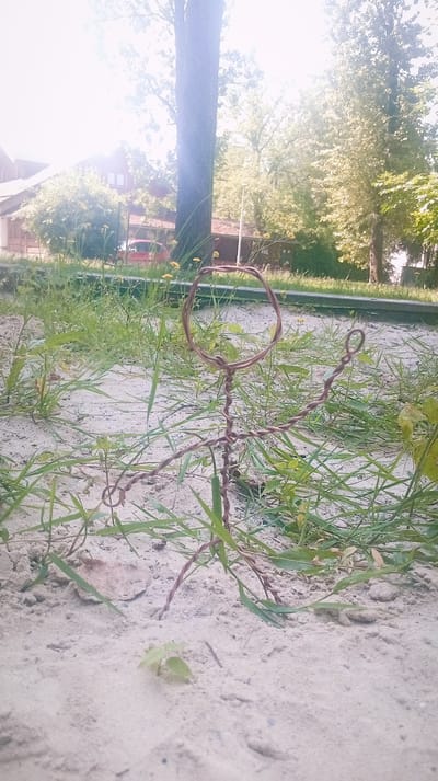 The image depicts a small, wire-sculpted figure resembling a person, made of brown wire, standing in sandy ground with sparse green grass and weeds. The figure has a circular head and a twisted body with outstretched arms. In the background, there is a blurred view of trees, buildings, and a red car. The lighting suggests it is daytime with bright sunlight. This scene is located in the city of Karviná, Czechia, specifically in a park or an open area with sand and vegetation.