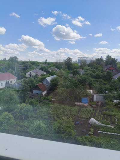 This is an elevated, eye-level, medium shot capturing a residential area with lush greenery under a partly cloudy blue sky. The scene appears to be a suburban or rural landscape, possibly within the city of Petrovsk, Russia, characterized by single-family homes, gardens, and abundant trees.
In the foreground, a white ledge or windowsill partially obstructs the bottom of the image. The middle ground is densely populated with a variety of structures and vegetation. Several houses are visible, varying in architectural style and roof color, with some featuring red or brown tiled roofs and others with lighter-colored walls. Some houses have visible chimneys. Surrounding the houses are numerous mature trees with full green canopies, creating a sense of established residential development.
Interspersed with the homes are garden plots. These gardens show signs of cultivation, with rows of plants, some covered with white protective sheeting, and dark soil. Various garden implements and structures, such as small sheds, fences, and what appear to be garden boxes or frames, are scattered throughout the area. A blue-sided structure is also visible, possibly a small shed or outbuilding.
In the background, the landscape continues with more trees and distant buildings. Notably, there are large industrial silos or storage tanks with a reflective surface visible in the far distance, suggesting a possible industrial zone or agricultural facility on the outskirts of the residential area. A cell tower is also present on the horizon.
The sky is a vibrant blue with scattered white cumulus clouds. The overall lighting suggests it is daytime, likely mid-morning or afternoon, with bright sunlight casting some shadows. There are no visible people or animals in the frame, and the scene conveys a sense of quiet, everyday life in a vegetated community. The image's perspective suggests it was taken from a higher vantage point, such as a balcony or upper floor of a building overlooking the neighborhood.