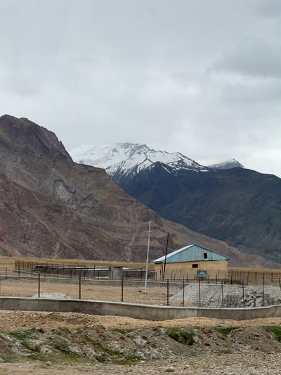 The image shows a building with a light blue roof in Zangla, India, set against a backdrop of imposing mountains, some of which are capped with snow. A wire fence encloses the area around the building, which appears to be a storage shed or small agricultural structure. The foreground is characterized by rough terrain with scattered rocks and sparse vegetation. The sky is overcast, suggesting a cloudy day. The scene is a rural, high-altitude landscape.