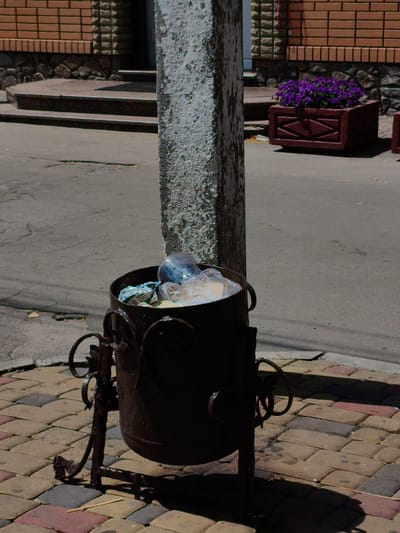 The image depicts an outdoor scene on a sunny day. In the foreground, a dark, weathered metal trash can with ornate wrought-iron legs stands on a multi-colored paved walkway. The trash can is partially filled with plastic bottles and bags. Behind the trash can, a rough, textured concrete pillar is visible. The walkway transitions to a grey asphalt surface, which appears slightly cracked. In the background, a building with a brick facade and stone foundation is present, with a set of stairs leading up to an entrance. To the right of the stairs, a dark red planter box filled with vibrant purple flowers adds a splash of color to the scene. This image does not provide any specific indicators of the location being in Velyka Bahachka, Ukraine. There are no people or animals visible in the frame.