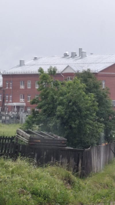 The image shows a brick building with a metal roof in the city of Yurla, Russia. The building has multiple windows and several vents on the roof. In front of the building, there are several large green trees. In the foreground, there is a dilapidated wooden fence with some wooden beams stacked behind it. The area is overgrown with grass and weeds. The sky is overcast.