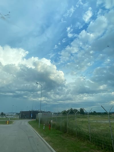 This media file shows a view from a vehicle moving along a paved road. The road appears to be on the outskirts of an industrial or commercial area, with a large, dark building on the left and a fenced-off grassy area with trees on the right. The fence is a chain-link fence with angled supports at the top. There are several streetlights visible along the road. In the distance, more buildings and parked cars can be seen. The sky is filled with dramatic clouds, with patches of blue sky visible. The clouds range from bright white cumulus to darker, more textured formations, suggesting a dynamic weather system.
Several small, dark objects are scattered across the sky, some of which are difficult to identify. They appear to be at a significant distance. One object in the upper portion of the sky seems to be a kite or a similar airborne recreational item, with a distinct shape suggesting a person or figure attached.
Red fire hydrants are visible next to the fence. A blue arrow sign is mounted on the building.
The lighting suggests it is daytime, likely mid-morning or afternoon, given the brightness of the sky and the shadows. The weather appears to be partly cloudy, with potential for changing conditions. The setting appears to be a modern infrastructure area, possibly near an airport or a large facility, given the fencing and the open, undeveloped land beyond. The overall mood is neutral, capturing a snapshot of movement through a functional landscape under a dynamic sky.