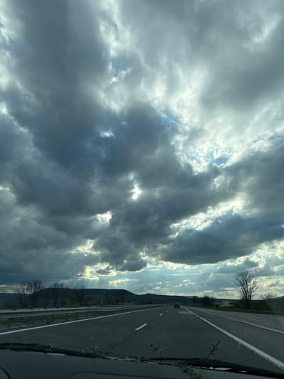 The image depicts a road scene in Zlatina, Bulgaria. A two-lane asphalt road with a white line dividing the lanes is visible. The road is adjacent to a grassy area and a mountain range can be seen in the background. The sky is filled with heavy, dark clouds, with some sunlight breaking through. A car is visible on the road ahead. The photograph appears to have been taken from inside a moving vehicle.