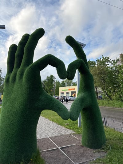 The image shows a large-scale public art installation of two hands made of grass, with the fingertips touching to form a heart shape. The hands are placed outdoors, likely in a park or pedestrian area, near the town of Ulyanovka, Russia. In the background, there's a yellow building with what appears to be a shop or office front, visible through the "heart" formed by the hands. Several people are standing outside this building, suggesting pedestrian activity. The environment includes a paved walkway, grass, trees, and a partly cloudy sky. A road is adjacent to the park.