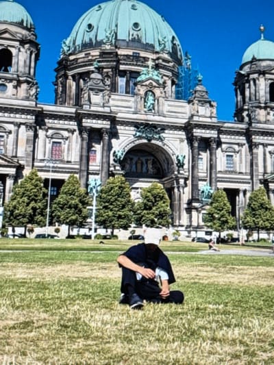 The Berlin Cathedral stands tall with its ornate domes and detailed architecture, set against a bright blue sky. A person dressed in black is sitting on the grass in front of the building. Several trees are aligned in front of the cathedral. People walk by in the background.