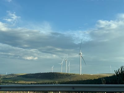 The image captures a landscape featuring a wind farm with multiple wind turbines situated on rolling hills under a partially cloudy blue sky. The foreground shows a grey metal railing and dry grass, suggesting a roadside view. The wind turbines are white, tall structures with rotating blades, positioned at various distances across the hills. The landscape is predominantly green and brown, with some scattered trees forming a dark band across the mid-ground. The sky exhibits a mix of clear blue and scattered grey clouds, with the sun illuminating parts of the scene. There are no people visible in the image. The scene depicts a rural or semi-rural environment, likely in a region with wind energy infrastructure. The location is identified as Urbnisi, Georgia.