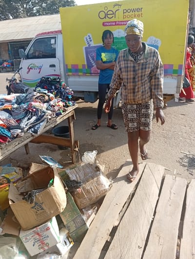 The scene depicts an outdoor market environment in Jebbu Miango, Nigeria, under bright daylight. In the foreground, a woman with a dark complexion walks towards the right side of the frame, wearing a light-colored patterned headwrap, a tan and dark brown plaid jacket, black and white patterned shorts featuring "CHANEL" logos, and light-colored flip-flops. Her arms are slightly bent, hands open.
Behind her, in the mid-ground, another woman with dark braided hair stands with her arms crossed, facing forward. She wears a blue t-shirt with yellow text/logo, black leggings, and black flip-flops.
To the left of the frame, a wooden market stall displays a large, disorganized pile of folded and stacked apparel items in various colors and patterns. Behind this stall is a white Godrej light-duty truck with a yellow cargo box bearing "Godrej aer power pocket Home Fresh" branding. A motorcycle is partially visible behind the stall.
On the ground in the lower foreground and mid-ground, a collection of cardboard boxes, some broken or flattened, are piled alongside plastic bags and other debris. Some boxes display brand names like "RODIS." To the right of these boxes, several rough-hewn wooden planks lie on the ground, forming a makeshift pathway or surface. In the background, partially visible structures and foliage can be discerned. The ground consists of compacted dirt or gravel.