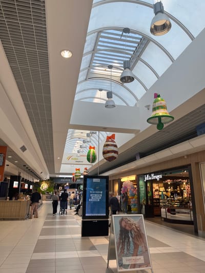 The image shows the interior of a shopping mall in Saint-Brice-Courcelles, France, located at coordinates 49.283, 4. The mall has a vaulted ceiling with a large skylight. There are multiple hanging light fixtures and festive decorations, including bells and Easter eggs. Several people are walking through the mall, passing various stores and kiosks. There is a large digital display on the left side of the image displaying a message in French. In the foreground, there is an A-frame sign advertising a brand called Saint Algue. On the right side of the image, there is a store called Jeff & Bruges.