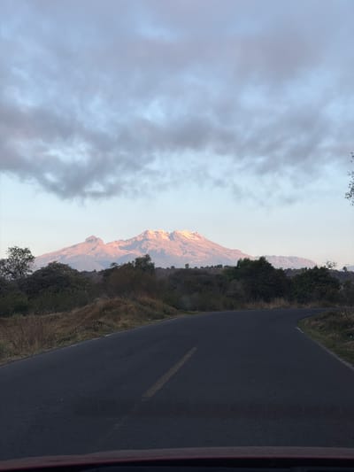 The image shows a road next to a mountain in Santiago Xalitzintla, Mexico. The road is asphalt and has a yellow line. The mountain is rocky and covered with vegetation. The sky is cloudy.