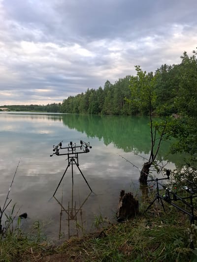 The image shows a serene lake scene in Nová Ves nad Lužnicí, Czechia. The foreground is occupied by a fishing setup with a tripod rod holder holding multiple fishing rods, positioned on the grassy bank of a tranquil, greenish-blue lake. The water reflects the overcast sky, which is filled with various shades of grey clouds. Dense green trees line the opposite bank of the lake, creating a lush backdrop. A single tree trunk stands in the water near the bank, with some fishing rods leaning against it. The shoreline in the foreground is covered with grass and some scattered debris. The overall atmosphere is one of quiet and anticipation, suggesting a fishing excursion.