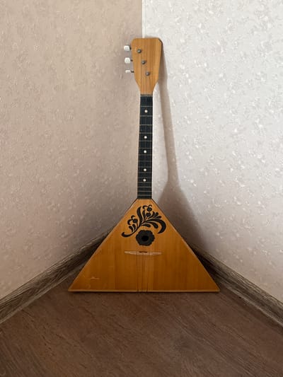 A wooden balalaika leans against a corner where two walls meet. The wall on the left has a light pink floral wallpaper, and the wall on the right has a white floral wallpaper. The floor is made of wooden planks. The balalaika is light brown with a black decorative element on its triangular body. The neck of the balalaika is black with white dots marking the frets. The top of the neck has three tuning pegs. There is no indication of this being located in Volkhov, Russia.