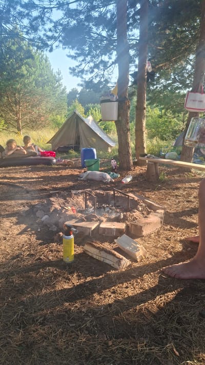The image depicts a rustic campsite scene, likely in a wooded area of Навасвержанскі сельскі Савет, Belarus. In the foreground, a makeshift campfire pit is visible, with embers and ash. A yellow aerosol can, possibly a lighter or fuel source, rests on the ground near the pit, alongside several pieces of firewood.
Further back, two children are lying on an inflatable mattress, appearing relaxed. A large, khaki-colored tent is pitched behind them. To the right of the tent, a blue plastic barrel and a green bucket are present, along with various other items suggesting a camping trip.
In the mid-ground, a white pig carcass is lying on the ground, possibly prepared for cooking. A wooden bench is situated to the right, with a glass of liquid resting on it. Two trees are prominent in the background, one with a white bucket and a bag attached to it, and another with a red and white bag that has "CVMARTANNI" printed on it. The overall environment suggests an outdoor recreational setting with elements of a temporary living space.