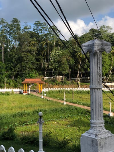 A serene landscape scene in Pilana, Sri Lanka, features a paved pathway leading through a lush, green field towards a traditional orange-roofed pavilion supported by yellow pillars. The pathway is lined with decorative white posts. In the foreground, a weathered, fluted stone column with Corinthian-style detailing stands prominently, partially obscuring the view. Overhead, thick black electrical wires crisscross the sky. The background is a dense forest of various trees, including palms, reaching up towards a partly cloudy blue sky. The field itself contains patches of water with lily pads and occasional pink flowers, suggesting a wetland area. Two individuals are visible on bicycles on the pathway, heading towards the pavilion, and another person is standing under the pavilion.