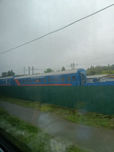 In Грабченки, Russia, a blue train with an orange stripe runs along a track, partially obscured by a green fence. The train has several windows and visible text that reads "ВОССТАНОВИТЕЛЬНЫЙ ПОЕЗД 3193". In the background, there are trees and industrial buildings. The sky is overcast and appears foggy or hazy. A dirt path with grass lines the side of the track. The image seems to have been taken from inside a moving vehicle, possibly another train, due to the motion blur.